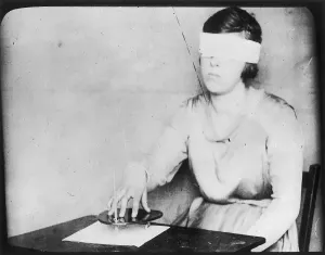 Prints of A girl, blindfolded, uses the planchette with a pencil fixed in it, hoping to receive by automatic writing a message from the spirits in the beyond Harry Price Library Prints: Blindfolded Girl Uses Planchette Print