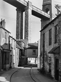 Prints of Street scene in Saltash, Cornwall, with Brunels Royal Albert Bridge towering above, crossing the River Tamar between Plymouth, Devon, and Saltash, Cornwall. Date: 1960s England Prints: Royal Albert Bridge Print, Saltash Cornwall 1960s