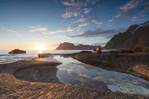 Pink clouds and midnight sun are reflected in the blue sea framed by rocky peaks Pink clouds and midnight sun are reflected in the blue sea framed by rocky peaks