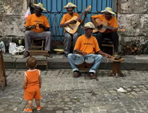 Play Collection: Street band wearing orange shirts playing music on the pavement watched by toddler wearing orange