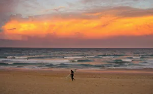 A surfer carrying his board walks out of the ocean as a cloud is lit by the setting A surfer carrying his board walks out of the ocean as a cloud is lit by the setting