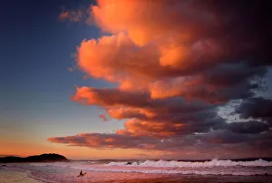 Surfer holding her board wades through the surf as clouds above are lit by the setting Surfer holding her board wades through the surf as clouds above are lit by the setting
