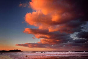 Wade Gallery: Surfer holding her board wades through the surf as clouds above are lit by the setting