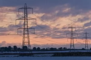 Electricity Collection: Electricity transmission pylons and overhead wires, crossing over snow covered farmland at sunset