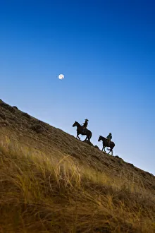 Two wranglers riding horses up a hill with full moon in backround at blue hour Two wranglers riding horses up a hill with full moon in backround at blue hour