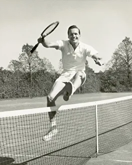 Man holding tennis racquet leaping over tennis net, 1950-60s, black and white Man holding tennis racquet leaping over tennis net, 1950-60s, black and white