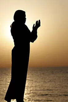 Silhouette of muslim woman in abaya praying with her hands up in air at sunset