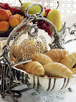 Antique silver hinged bread basket with croissants in front of a bowl of fruit on a Antique silver hinged bread basket with croissants in front of a bowl of fruit on a