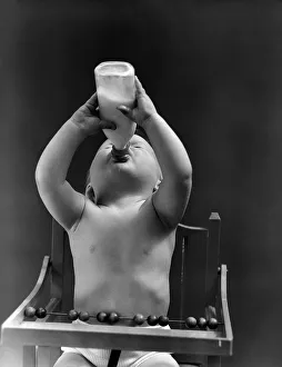 Baby sitting in high chair, head tilted back, drinking from milk bottle. (Photo by H Baby sitting in high chair, head tilted back, drinking from milk bottle. (Photo by H