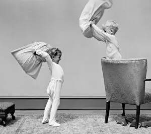 Images Dated 30th June 2008: Boy and girl having a pillow fight, boy standing on chair swinging pillow, girl on floor