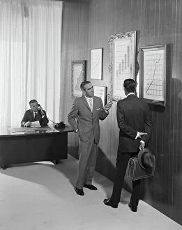 Images Dated 30th July 2011: Two businessman discussing at bar chart while another man using telephone in background