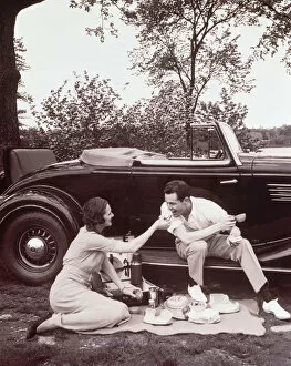 Couple picnicking, man sitting on car runningboard (B&W sepia tone)