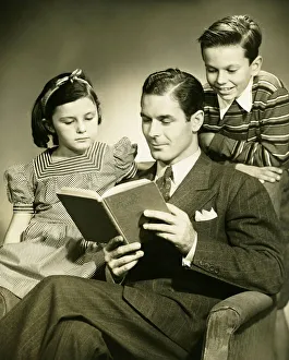 Father reading to son (4-5) and daughter ( 6-7) in studio, (B&W), portrait Father reading to son (4-5) and daughter ( 6-7) in studio, (B&W), portrait