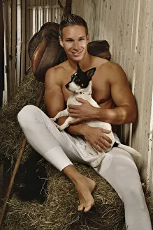 Man with a naked torso wearing long underwear with a dog in a horse barn sitting on bales Man with a naked torso wearing long underwear with a dog in a horse barn sitting on bales