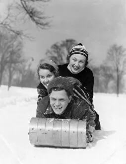 Man And Two Women Wearing Wool Hats And Winter Clothing Tobogganing In Snow With Tree