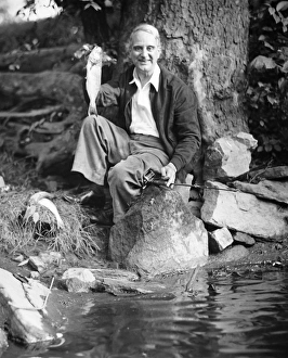 Senior man sitting by water, holding up fish (B&W), portrait Senior man sitting by water, holding up fish (B&W), portrait