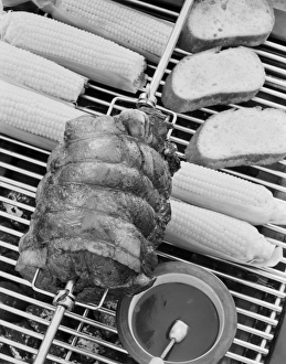 Spit roasting of ham on barbecue grill with corn cobs, close-up Spit roasting of ham on barbecue grill with corn cobs, close-up