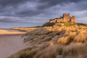 Bamburgh Castle and sand dunes in warm, late evening light with stormy evening sky Bamburgh Castle and sand dunes in warm, late evening light with stormy evening sky