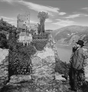 Historic image in black and white of a man standing and smoking a pipe with a view of Rheinstein Castle Historic image in black and white of a man standing and smoking a pipe with a view of Rheinstein Castle