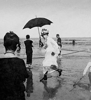 Historic image in black and white of a Victorian lady with umbrella running in the surf on the beach
