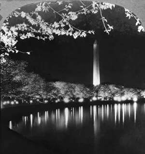 Historic image in black and white of the Washington Monument illuminated at nighttime and reflected in a river