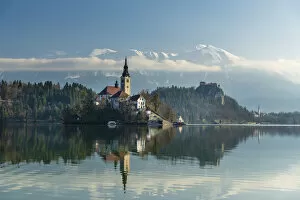 Looking across the calm waters of Lake Bled at dawn to the church on Lake Bled Island and Bled Castle behind; Bled