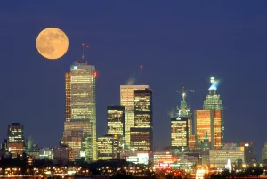 Full Moon In An Orange Hue Over A City Skyline Illuminated At Nighttime Full Moon In An Orange Hue Over A City Skyline Illuminated At Nighttime
