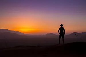 Images Dated 26th December 2015: Silhouette of man wearing a cowboy hat looks out at the glowing landscape and sky at sunset, Namibia