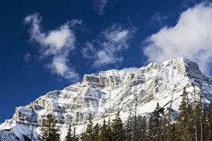 Snow Covered Mountain Peak With Evergreen Trees With Blue Sky And Clouds; Banff, Alberta, Canada Snow Covered Mountain Peak With Evergreen Trees With Blue Sky And Clouds; Banff, Alberta, Canada