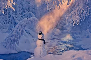 Snowman Standing Next To A Stream With Sunrays Shining Through Fog And Hoar Frosted Trees In The Background