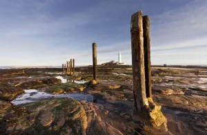 Wooden Posts At The Water's Edge With A Lighthouse In The Distance At Whitley Bay; Northumberland, England Wooden Posts At The Water's Edge With A Lighthouse In The Distance At Whitley Bay; Northumberland, England