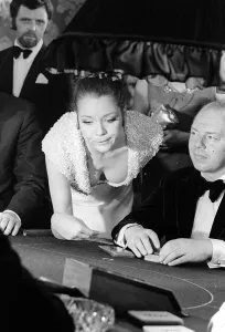 Core26 Gallery: Diana Rigg in a low cut dress leans across the Black Jack table in a casino showing off
