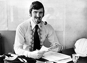 Jimmy Hill sitting at his desk smoking a pipe, dressed in stylish 1970s clothing