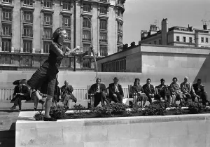 Prints of Fashions taken during London Fashion Week 1964 Model wearing a tartan skirt and waistcoat with a bow and arrow - archery - as a row of men sitting down on a bench look on Event Prints: London Fashion Week 1964 Print: Tartan Skirt and Waistcoat