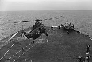 A Royal Navy Westland Sea King hovers over the deck as a Buccaneer with folded wings sits