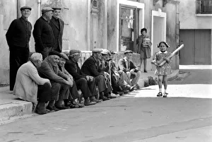 Walking past the old men of Poussan is 6 year old Nathalie Rustan carrying her French Walking past the old men of Poussan is 6 year old Nathalie Rustan carrying her French