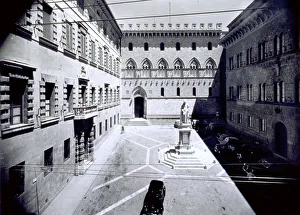 Piazza Salimbeni in Siena. The buildings on three sides of the square are Palazzo Spannocchi on the right, Palazzo Tanucci on the left, and Palazzo Salimbeni at the back. The statue of Sallustio Bandini stands in the center of the square. A few cars are parked in the square