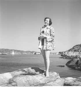 Retro Swimwear Fashion Prints: 1950s summer fashion. A young woman in a bikini with a light short coat pictured standing on a cliff on the coast